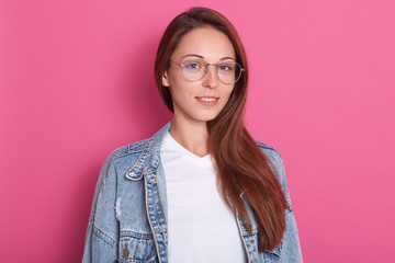 Fototapeta premium Indoor shot of pleasant looking woman, smiles positively, wearing optical glasses, white t shirt and denim jacket, being in hight spirt, model posing isolated over pink background. Copy space.