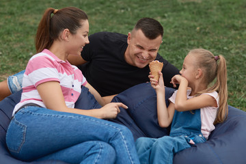 Outdoor shot of happy family enjoy spending time together in park. Doughter gives father to taste her ice cream, family having fun together in open air, looks happy. Childhood and happyness concept.