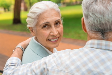 Smiling old woman with look at camera at park with her husband with view from back