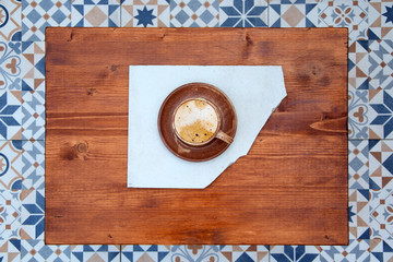 Cup of cappuccino on wooden table. Still life detail in home interior. Wooden table with cup of coffee on it and colorful tiles top view