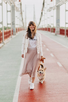 Attractive young woman walking corgi outdoors. Positive female with glad expression with her dog being satisfied outdoor