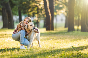 full length view of beautiful young girl in casual clothes hugging golden retriever while sitting on meadow in sunlight and looking at camera