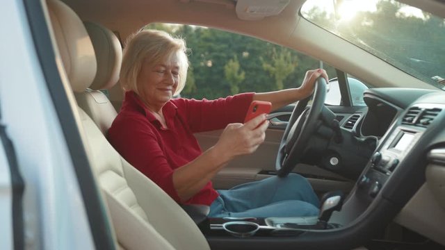 At sunset senior woman sits in car use red cell phone smile happy businesswoman blonde people smartphone cellphone communication internet search lady mobile bench browse network online slow motion