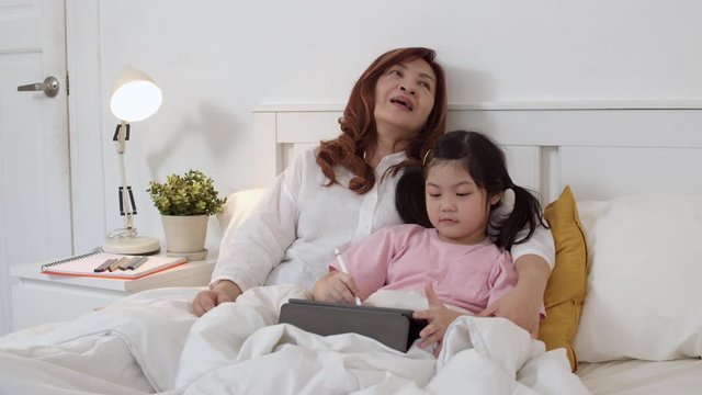Asian grandmother using tablet read fairy tales to granddaughter at home. Senior Chinese, grandma happy relax young girl before bedtime lying on bed in bedroom at home at night concept. Slow motion.