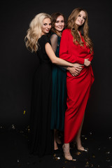 Studio full length portrait of three stunning young ladies in evening gowns posing for camera. Black background. New Year evening celebration birthday concept.