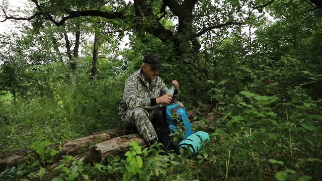 handsome Caucasian traveler in camouflage clothes wearing a cap and a backpack, sitting resting on a log in a dense green forest, opens a bottle of water and drinks, close-up