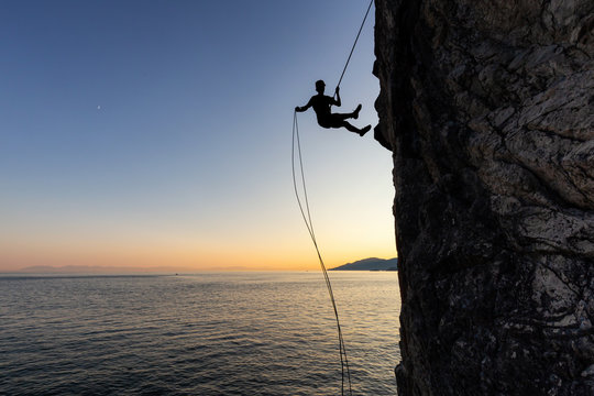 Silhouette of a Unrecognizable man rappelling down a steep cliff on the rocly ocean coast during a sunny summer sunset. Taken in Lighthouse Park, West Vancouver, British Columbia, Canada.