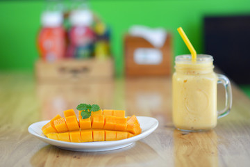 Mango smoothie in tall glass with plate of diced mangoes on wooden table.