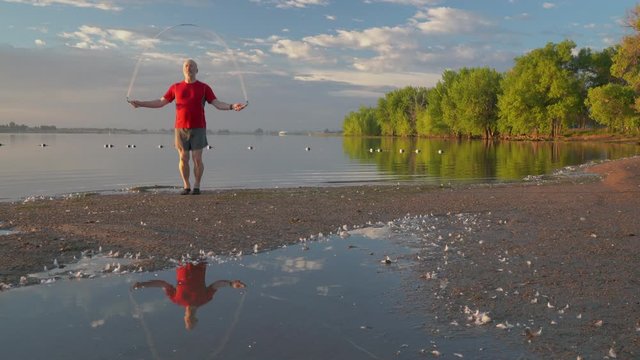 Senior man is exercising with a heavy  fitness jump rope on a sandy lake beach - summer morning in Boyd Lake State Park in northern Colorado