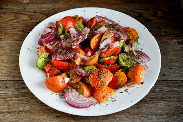 Summer salad of tomatoes, cucumbers, spices and herbs in a white plate on a wooden table.