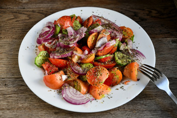 Summer salad of tomatoes, cucumbers, spices and herbs in a white plate on a wooden table.