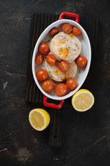 Baked hake medallions with cherry tomatoes and lemon, top view on a dark brown stone background, vertical shot