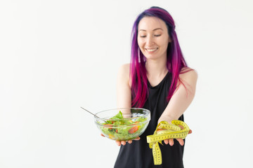 Slim young asian girl with colored hair is holds a measuring tape and holding a light vegetable salad in her hands posing on a white background. Healthy eating concept. Copyspace.