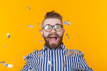 Joyful young hipster male in a glasses is laughing happily among flying confetti on a yellow background. The concept of a mega party and holiday.