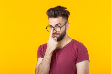 Pensive young hipster man with a beard holding his glasses while standing against a yellow...