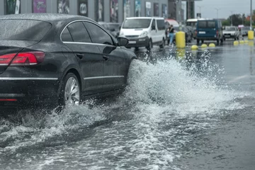 Foto op Aluminium  Driving car on flooded road during flood caused by torrential rains. Cars float on water, flooding streets. Splash on the car. Flooded city road with a large puddle © Aleksandr Lesik