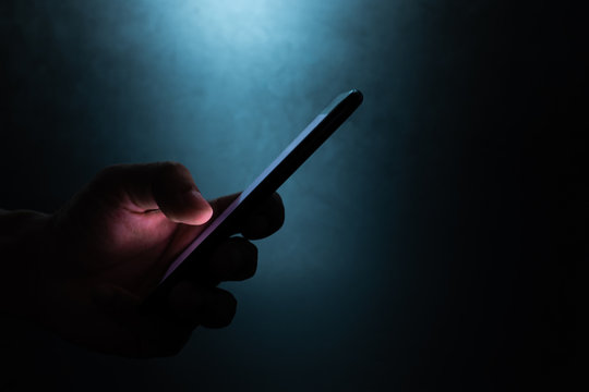 silhouette hand holding and touching a mobile phone screen in the dark darkness against dark blue background