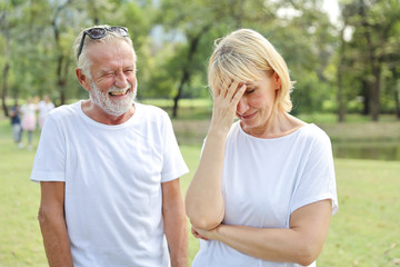 elderly wife is upset and husband is looking at her while laughing and try to soothe her a the same time in park with green trees during summer time