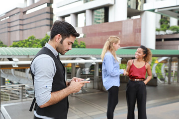 side view successful caucasian businessman in black suite who standing in downtown, he using cell phone with his colleagues blurred background