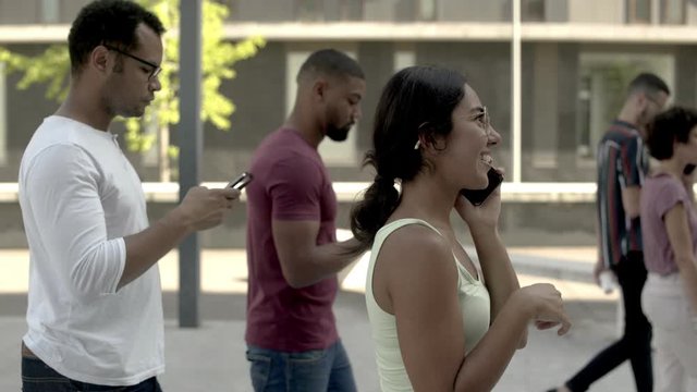 Side View Of Excited Woman Talking On Phone In Crowd. Group Of Young People Walking On Street And Talking Or Texting On Smartphone. Communication And Technology Concept