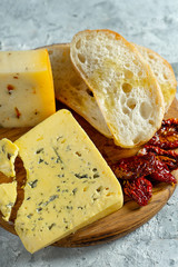 Different types of cheese on wooden cutting board. blue cheese with ciabatta and sun-dried tomatoes on grey background. Italian cuisine dinner. Copy space. Soft focus, top view