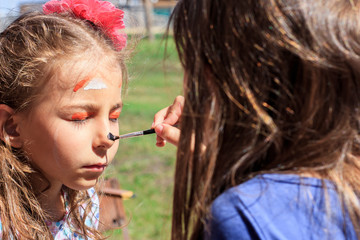 A girl paints a fox on her sister's face with watercolors, depicting face painting