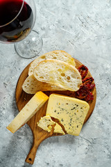 Different types of cheese on wooden cutting board. blue cheese with ciabatta and sun-dried tomatoes and glass of wine on grey background. Italian cuisine dinner. Copy space. Soft focus, top view