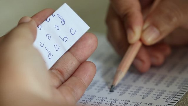 Education concept. Student's hands filling in standardized test form with black pencil drawing selected choice on answer sheets and cheating in test with answer piece of paper in hand. 