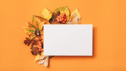 Autumn holiday invitation. White mockup paper sheet with foliage decoration on orange background. Copy space.