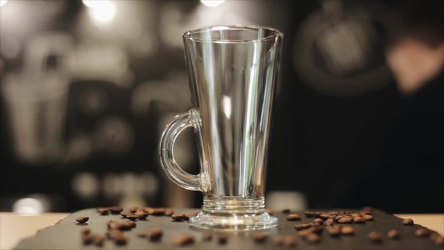 A close-up of an empty glass cup for latte standing on a bar counter with some coffee beans on it