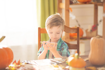Happy Halloween concept. Cute little happy boy carving a pumpkin and decorated cookies for halloween on a table indoors.