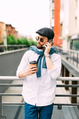 Business talk on a go. Cheerful young man in sunglasses holding coffee cup and talking on mobile phone with smile while walking outdoors