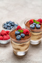 Classic tiramisu dessert with blueberries and raspberries in a glass and bowls with berries on concrete background
