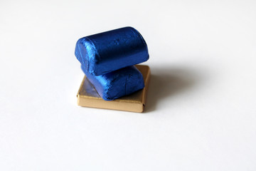 A pile of chocolate candies wrapped in blue and golden foil isolated on white surface. Template for greeting card, poster or invitation. Sweets scattered on white smooth table. Backdrop or background