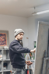 Young, positive artist works, paints a picture of oil paints in a cozy studio. Artist with a canvas on an easel places an oil paint on a palette. Process of creating a picture. Copyspace