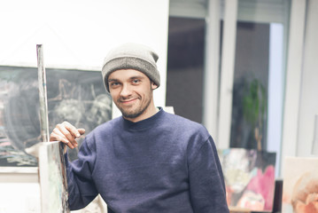 Portrait of a close-up of a happy artist in a hat and a blue sweater, looking into the camera and smiling. Creative man in a creative studio.