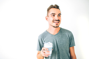 Happy young guy in grey t-shirt holding a cup of coffee on white isolated background. Looking away smiling.