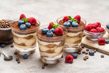 Classic tiramisu dessert with blueberries and raspberries in a glass and bowls with berries on concrete background