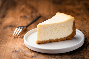 Cheesecake slice, New York style classical cheese cake on wooden background. Slice of tasty cake on white plate served with dessert fork - obrazy, fototapety, plakaty