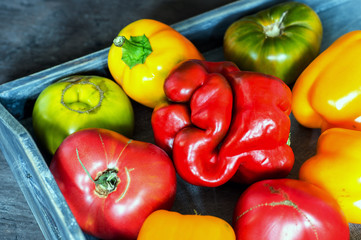 Imperfect natural peppers and tomatoes on an old wooden tray on a dark background. Healthy eating concept.