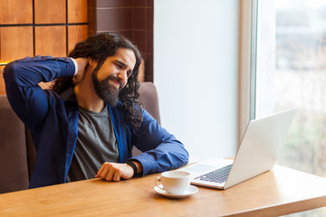 Portrait of tired handsome young adult man freelancer in casual style sitting in cafe with laptop, holding his neck after hard overtime work and make massage of neck pain. Indoor, lifestyle concept