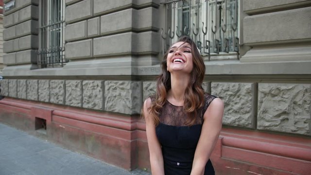 beautiful girl, brunette, in a dark blue long dress with lace, with curls and bright makeup, crown on her head, posing and smiling in front of the camera. is happening on the street near buildings