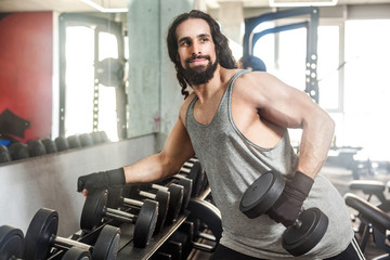 Fototapeta premium Portrait of confident young adult man handsome athlete with long curly hair working out in gym, standing, holding one dumbbell with raised arm, doing exercises for biceps. indoor, looking away, smile