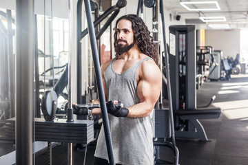 Fototapeta na wymiar Portrait of young adult sport athlete man with long curly hair training at gym alone, standing and lifting weights in the gym, doing exercises for biceps. Healthy lifestyle concept, indoor