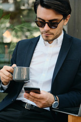 Young attractive bearded brunette man in white shirt,classic jacket and sunglasses holding cup of coffee in hands thoughtfully using cellphone on city street alone