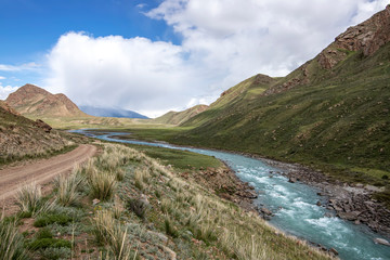 Fototapeta na wymiar View of the river flowing between green hills with grass and stones in the foreground Kyrgyzstan. Tien Shan