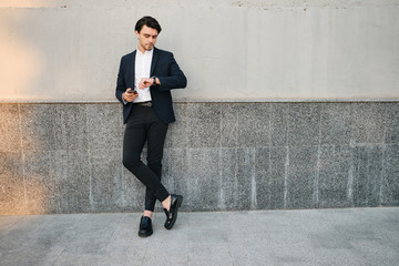 Young handsome bearded brunette man in white shirt and classic suit with wireless earphones holding cellphone in hand dreamily looking on wristwatch near gray wall on street alone