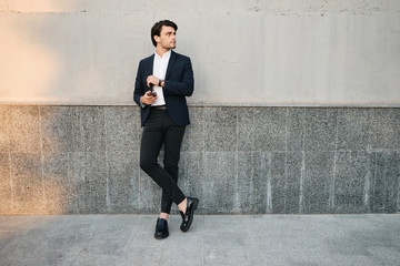 Young beautiful bearded brunette man in white shirt and classic suit with wireless earphones holding cellphone in hand dreamily looking aside while standing near wall on street