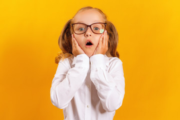 Portrait of a cheerful little girl on a yellow background. The child looks in surprise at the camera and holds her face in his hands. Education and school concept.