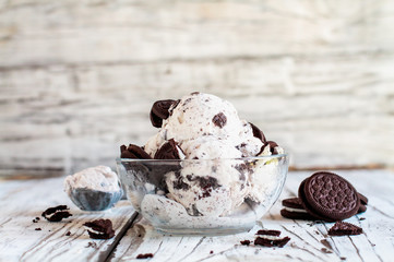 Clear glass bowl of cookies and cream ice cream. Selective focus with blurred background. Whole and crumbled cream filled cookies scattered about table. 
